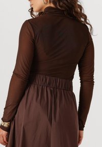Bruine langemouwtop met hoge kraag en doorschijnende textuur, gecombineerd met een bruine rok met elastische taille, die een gladde stof en een getailleerd ontwerp toont.