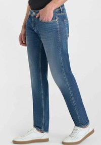 Jeans in denim blu slim-fit con una singola tasca frontale, cuciture in vista e una texture leggermente sbiadita, abbinati a sneakers bianche.