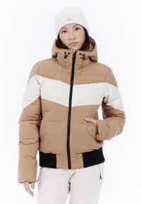 Hnedá puffa bunda s bielym šípkovým pruhom, nastaviteľnou kapucňou a čiernymi rebrovanými manžetami, doplnená svetlou čiapkou.