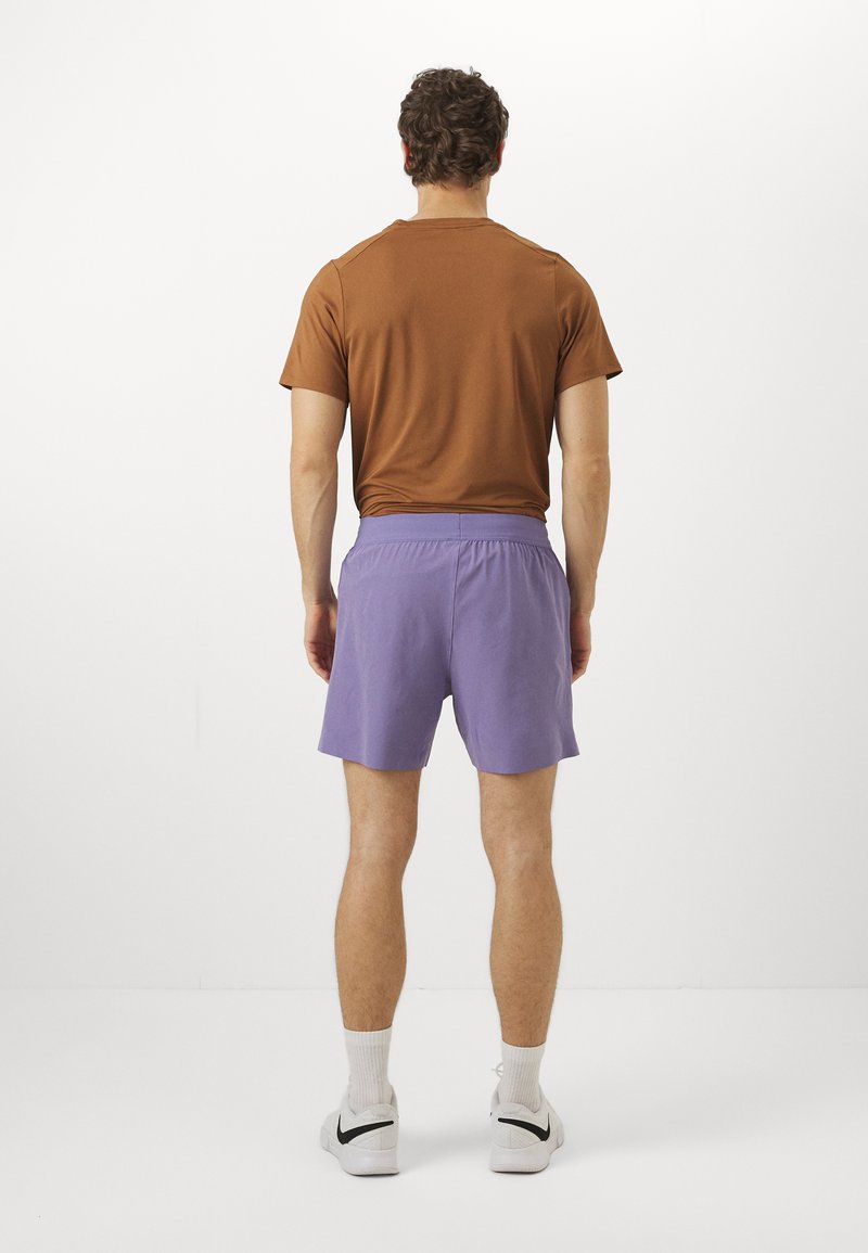 Braun kurzärmeliges Hemd und lila Shorts mit elastischem Bund. Das Model trägt weiße Sneakers mit schwarzen Akzenten und gerippten weißen Socken.