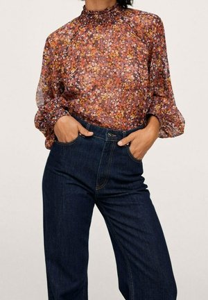 Woman wearing a floral long-sleeve blouse with a high ruffled neck tucked into dark blue high-waisted jeans, hands in pockets.