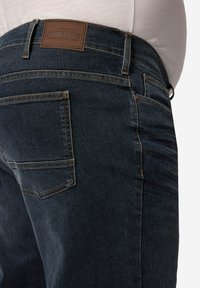 Jeans in denim blu scuro con cuciture arancioni, dotati di un'etichetta in pelle marrone sulla vita e un classico design a cinque tasche.