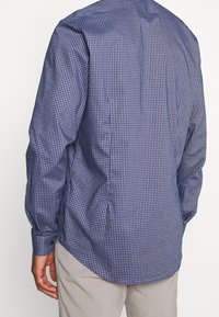 Långärmad skjorta i marinblå och vit rutigt mönster, med knäppte manschetter, rundad nederdel och slät textur. Bakåtvyn visar en skräddarsydd passform.