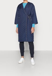 Persona che indossa un cappotto trapuntato blu navy sopra una maglietta a maniche lunghe blu brillante, pantaloni neri e sneakers bianche, in piedi su un pavimento chiaro.