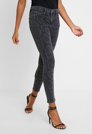 Person trägt schwarze Snake-Print Skinny-Jeans und schwarze Sandalen mit Absatz und Knöchelriemen, steht vor einem schlichten Hintergrund.