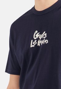 Camiseta de algodón azul marino con cuello redondo y mangas cortas; presenta texto blanco "Guess Los Angeles" en una fuente casual en la parte delantera.