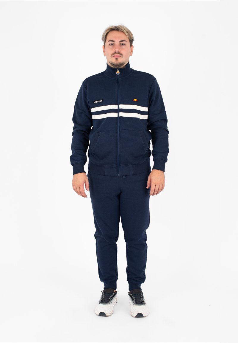 Completo tuta navy con giacca con zip e pantaloni coordinati, caratterizzato da strisce bianche sul petto, polsini a coste e tasche laterali.