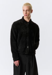 Μαύρο, φουντωτό μπουφάν με φερμουάρ και ψηλό γιακά, με απαλή υφή. Φοριέται με χαλαρό μαύρο παντελόνι, τονίζοντας μια άνετη εφαρμογή.