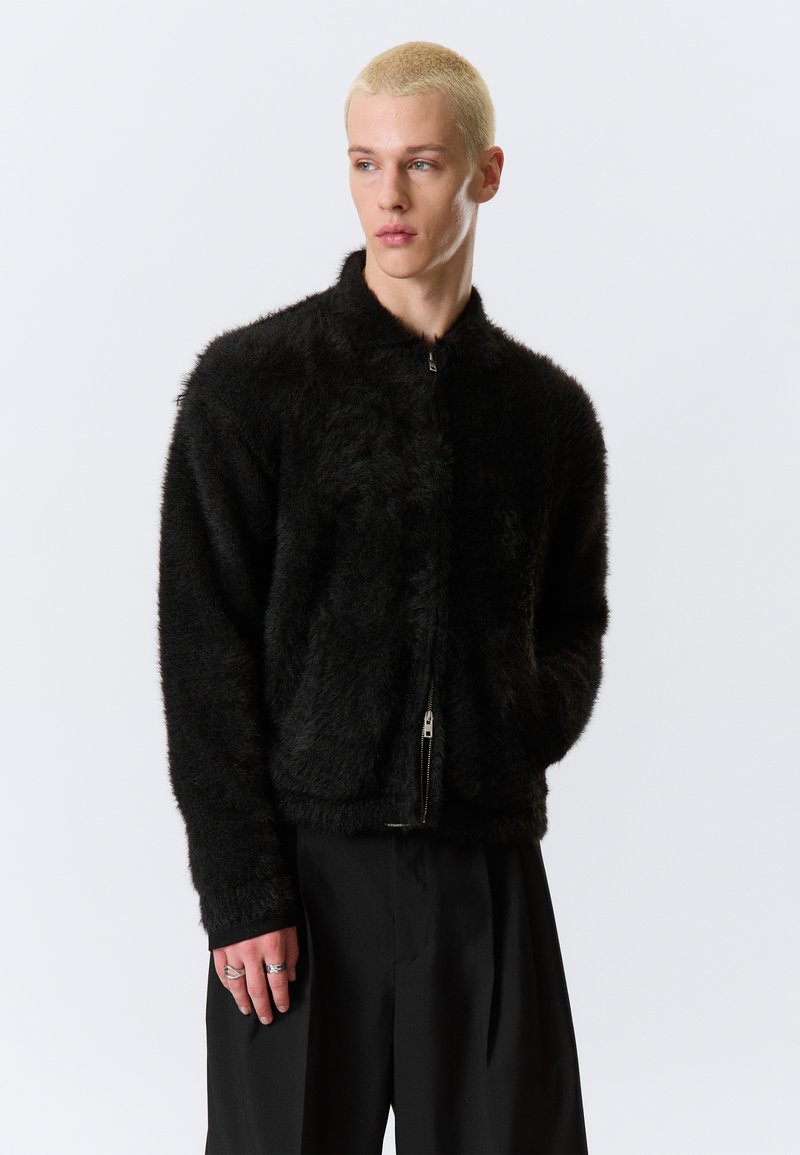 Μαύρο, φουντωτό μπουφάν με φερμουάρ και ψηλό γιακά, με απαλή υφή. Φοριέται με χαλαρό  μαύρο παντελόνι, τονίζοντας μια άνετη εφαρμογή.