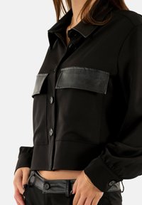 Femme portant une veste noire courte avec des rabats de poche en cuir et un pantalon noir assorti avec des détails de ceinture en cuir.