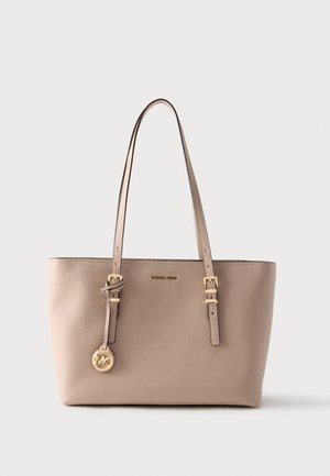 Béžová kabelka z hrubozrnné kůže s dvojitými ramenními popruhy, zlatými přezkami a kulatým zlatým přívěskem s logem MK zavěšeným na jednom popruhu.