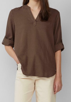 Frau trägt eine braune Bluse mit langen Ärmeln und aufgerollten Manschetten sowie cremefarbene Hose, steht mit einer Hand in der Tasche vor einem schlichten Hintergrund.