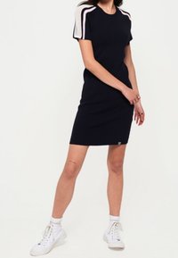 Abito in maglia navy con maniche corte, caratterizzato da dettagli a righe rosa chiaro e bianche sulle spalle. Indossato con sneakers bianche. Design semplice e lineare.