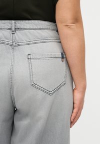 Prim-plan cu o persoană purtând blugi gri deschis cu un buzunar pe spate și o cămașă neagră, arătând partea laterală a brațului și mâinii lor.