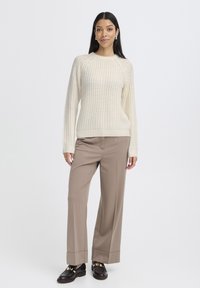 Cremig gestrickter Pullover mit Raglanärmeln, kombiniert mit beigen weit geschnittenen Hosen. Braune Schuhe mit goldenen Akzenten runden das Outfit ab.
