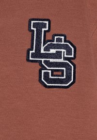 Brauner Baumwollstoff mit einem strukturierten, marineblauen und weißen gestickten "LS"-Logo. Das Design zeichnet sich durch eine weiche, glatte Textur aus.