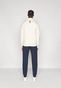 Λευκό φούτερ με ψηλό γιακά και μαύρη ρίγα στην πλάτη, σε συνδυασμό με ναυτικό μπλε παντελόνι φόρμας. Λευκά αθλητικά παπούτσια ολοκληρώνουν την εμφάνιση.