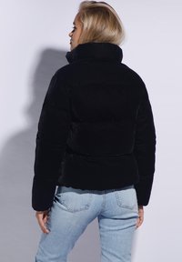 Sorte cropped pufferjakke med høj krave, blød tekstur, vandrette sømme og elastiske manchetter, parret med lyseblå jeans.