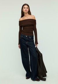 Ženska v rjavem off-shoulder dolgih rokavih topu in širokih temnih kavbojkah drži črn usnjen jopič pred enotno ozadje.