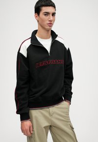 Μαύρο φούτερ με μανίκια και λαιμόκοψη τύπου ζιπ-ζακου, με κόκκινες και λευκές λεπτομέρειες, που περιλαμβάνει το κείμενο "LETS FRANS" με κόκκινη κέντηση. Ριμπ μανίκια και στρίφωμα, από μείγμα βαμβακιού.