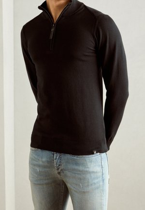 Pull noir à col zippé avec un design ajusté, des poignets et un ourlet côtelés. Porté par-dessus un jean déchiré bleu clair, mettant en valeur un tissu texturé.