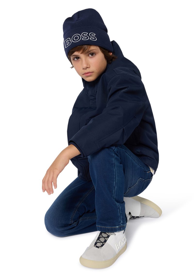 Doudoune marine, jeans bleus et baskets blanches avec des accents gris. Un enfant est en position à genoux, portant un bonnet en tricot marine assorti avec le texte "BOSS".