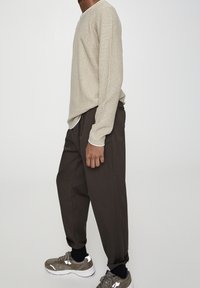 Pull en maille beige à manches longues, coupe décontractée, associé à un pantalon fuselé marron foncé. Baskets grises avec des accents blancs et chaussettes noires visibles.