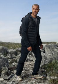 Padded black jacket over a gray shirt, paired with dark pants and multicolored hiking shoes. A backpack is worn on the back.
