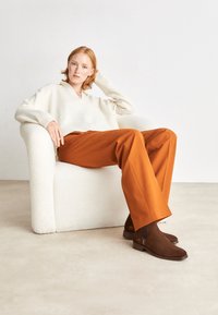 Pull en tricot crème avec un col en V, associé à un pantalon orange à jambes larges. Assis dans un fauteuil texturé blanc, avec des bottines marron.