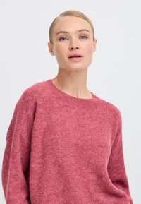 Maglione rosa a maglia con una texture morbida, scollatura rotonda e spalle calate. Il tessuto appare accogliente e leggermente peloso.
