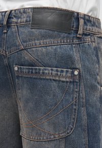 Modre kavbojke iz jeansa z teksturirano površino, z oranžnim šivom, detajlom zadnjega žepa in črno usnjeno etiketo na pasu.