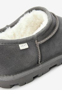 Grå sutsko med en blød, hvid pelsforing. Har en tekstureret overdel, et syet design og en gummisål med udtalt mønster. Etiket er synlig.
