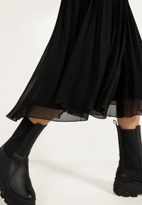 Gonna nera plissè con sovrapposizione trasparente, abbinata a stivali alla caviglia in pelle nera spessa con pannelli laterali elastici e suole testurizzate.