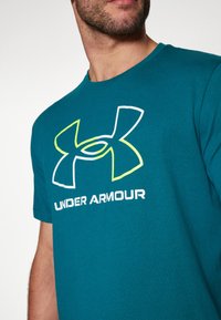 Teal kortærmet t-shirt lavet af bomuld, der har en stor hvid og neongrøn Under Armour-logo på tværs af brystet. Flad halsudskæring.