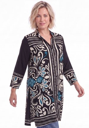 Blond vrouw die een lange zwarte tuniek draagt met witte en blauwe bloemmotieven, staand tegen een witte achtergrond.