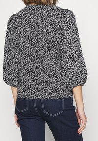 Blouse noire à motifs floraux avec manches bouffantes, ornée d'un motif floral blanc. Tissu lisse, ourlet arrondi et détail d'encolure froncée.