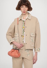 Beige denim jacket with floral embroidery on sleeves and pockets, paired with matching pants. Pink leather bag with a gathered design.