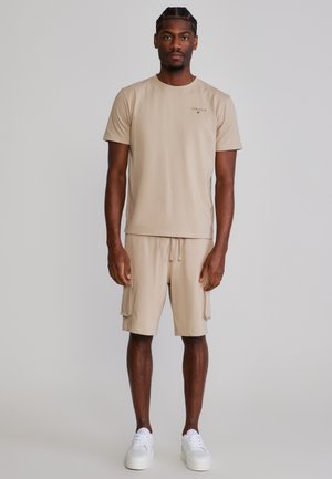 Muž v béžovej košeli s krátkym rukávom a zodpovedajúcich cargo šortkách s bielymi teniskami, stojaci pred jednoduchým sivým pozadím.