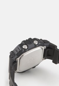 Montre en caoutchouc noir avec un boîtier métallique rectangulaire, deux boutons sur le côté et un dos en acier inoxydable avec des détails gravés.