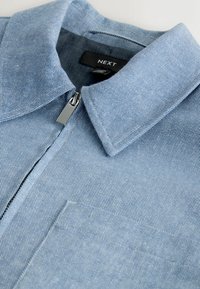 Chemise en denim bleu clair avec col pointu, fermeture éclair argentée et poche poitrine visible, portant l'étiquette de marque "NEXT" à l'intérieur du col.