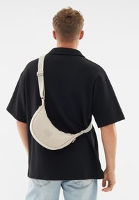 Borsa a spalla color crema con forma curva a mezzaluna, dotata di tracolla e di un sottile etichetta del marchio, indossata sopra una maglietta nera.