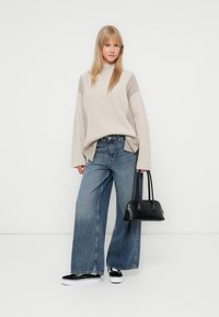 Pulover oversized bej, tricotat, cu mâneci largi, asociat cu blugi evazați de culoare albastru deschis. O geantă neagră și adidași negri completează ținuta.