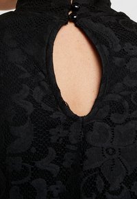 Svart spetsblus med hög krage, texturerat blommönster och nyckelhål bak med två svarta knappar. Slät tygfinish.