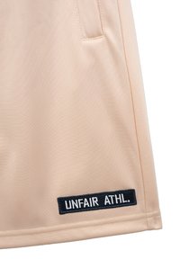 Jasnopomarańczowa spódnica sportowa, wykonana z gładkiego materiału o subtelnej teksturze. Posiada czarną haftowaną naszywkę z napisem "UNFAIR ATHL."