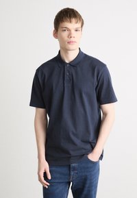 Polo de manga curta em algodão azul marinho, com colarinho clássico, pala de três botões e logótipos bordados discretos no peito.