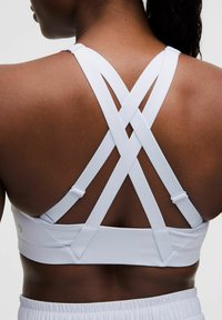 Sujeta de deporte blanca con tirantes cruzados, hecha de un material suave y elástico. Presenta tirantes ajustables y un diseño ajustado.