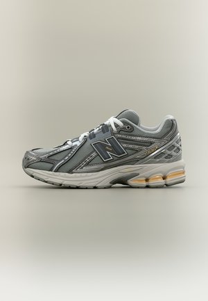 Ténis New Balance 1906R cinzento com atacadores brancos, parte superior em malha e couro, e sola acolchoada com inserções laranja semelhantes a gel.