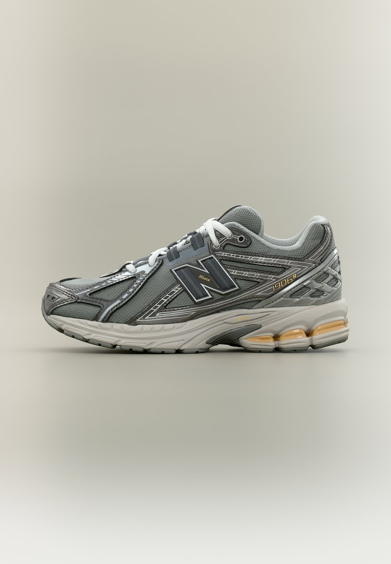 Γκρι αθλητικό παπούτσι New Balance 1906R με λευκά κορδόνια, επάνω μέρος από διχτυωτό ύφασμα και δέρμα, και ενισχυμένη σόλα με πορτοκαλί ένθετα που μοιάζουν με τζελ.