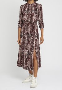 Maxiklänning i mönster med vågor i röd och beige, med hög halsringning, långa ärmar, cinchad midja och en sidoklyfta, i kombination med vita sneakers.