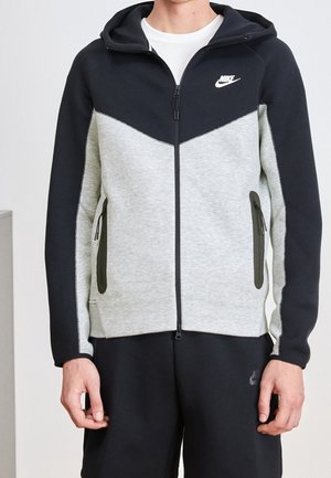 Mężczyzna ubrany w dwukolorową, czarno-szarą bluzę Nike na zamek z kieszeniami na zamek oraz czarne spodnie sportowe.